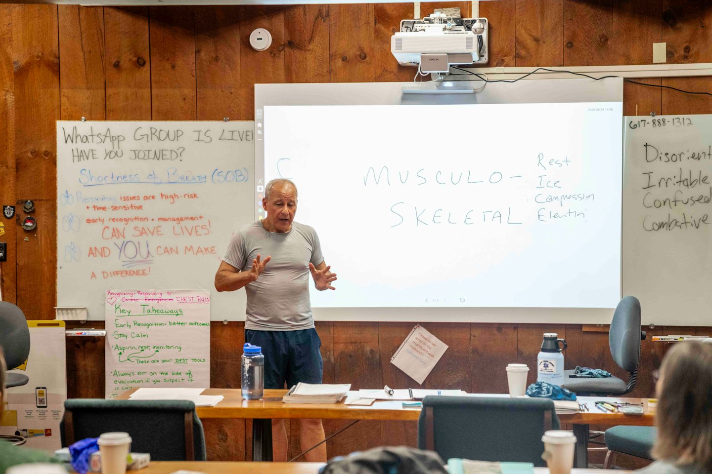 Un homme plus âgé enseigne à une classe en gesticulant devant des tableaux blancs remplis de notes.